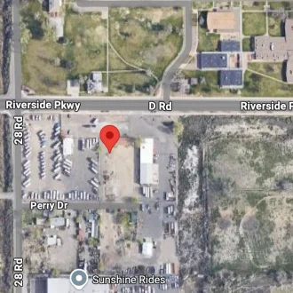A satellite view of an industrial area near the intersection of Riverside Parkway and 28 Rd, with a location pin marking a lot on Perry Dr. Residential and open areas surround the site.