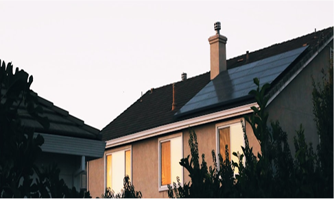 A house with solar panels installed on the roof, illuminated by the setting sun, framed by tree branches in the foreground.